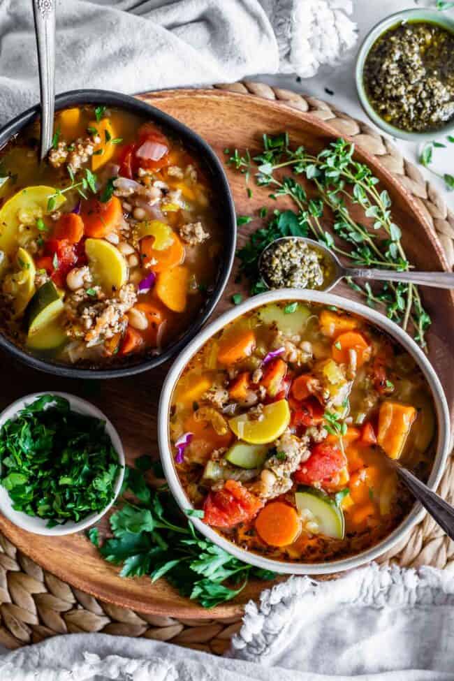 turkey vegetable soup in two bowls on a wooden charger plate with parsley and thyme garnish on the side.
