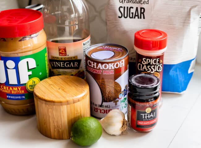 peanut butter, vinegar, salt, coconut milk, lime, garlic, red curry paste, sugar, on a white counter.