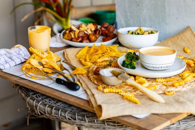 photo shoot set up: fries, chips, broccoli, cheese dip on a wooden board with brown table runner.
