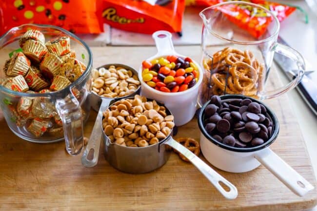 ingredients in measuring cups on a cutting board: Reese's cups, peanuts, peanut butter chips, Reese's pieces, chocolate chips.