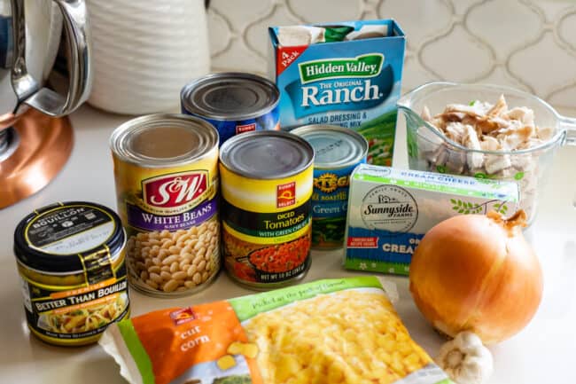 ingredients for white chicken chili recipe set up on a counter: canned beans, onion, corn, ranch mix, cream cheese, etc.