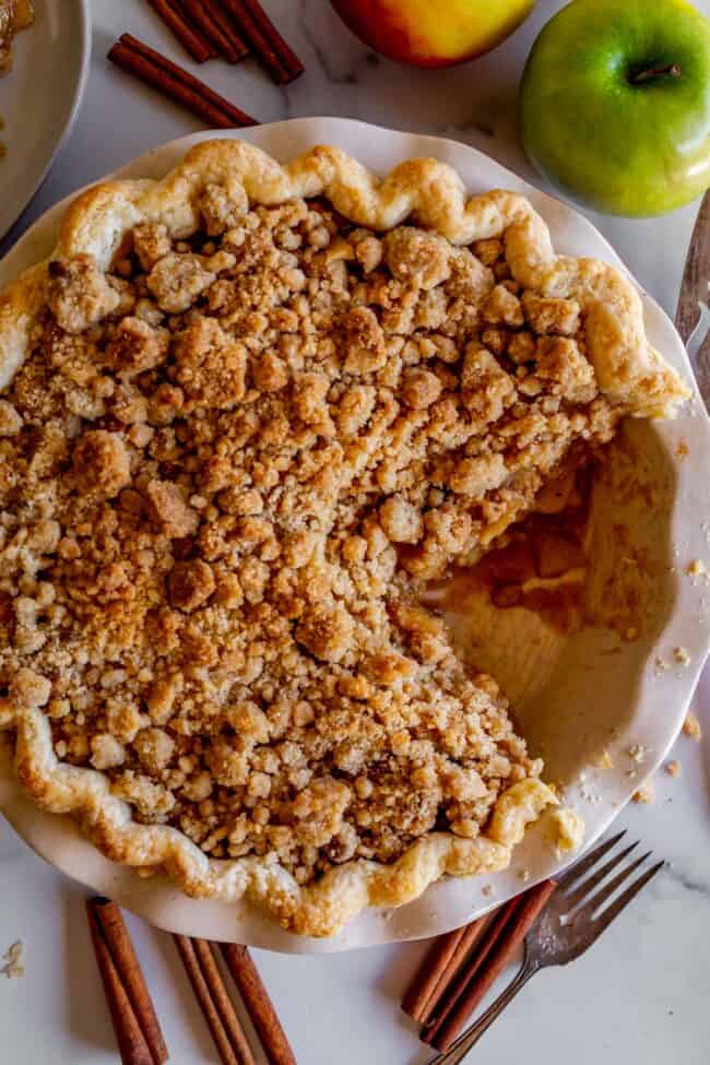 crumble top dutch apple pie in a white pie plate with a large slice cut out of it.