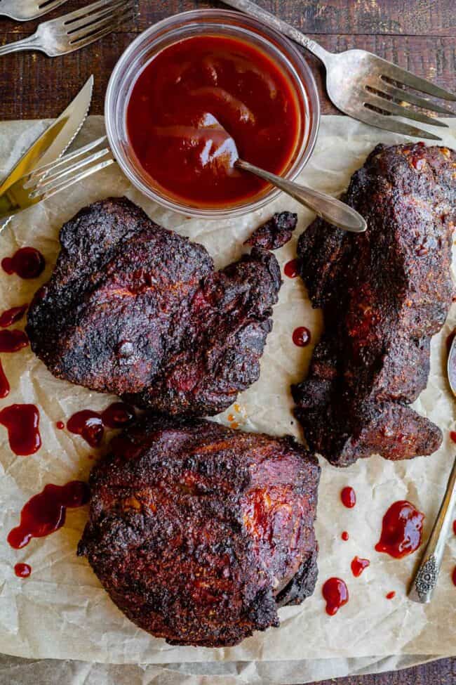 oven roasted pork shoulder in 3 pieces on parchment paper with bowl of bbq sauce.