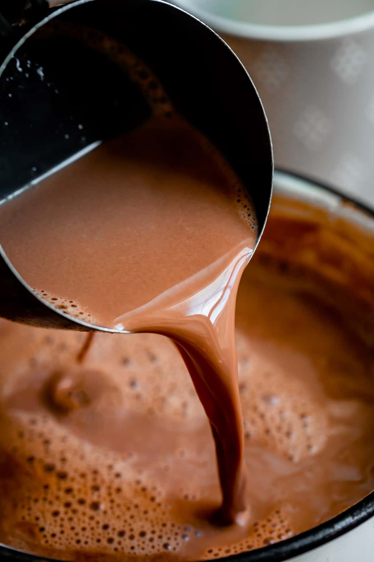 a black measuring cup pouring best hot chocolate into a white pot.