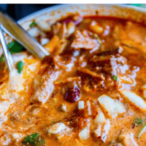 A close-up of a bowl of rich, hearty slow cooker chili topped with sour cream, shredded cheese, and herbs, with a spoon resting inside. This chili appears thick and flavorful, filled with chunks of meat and beans.