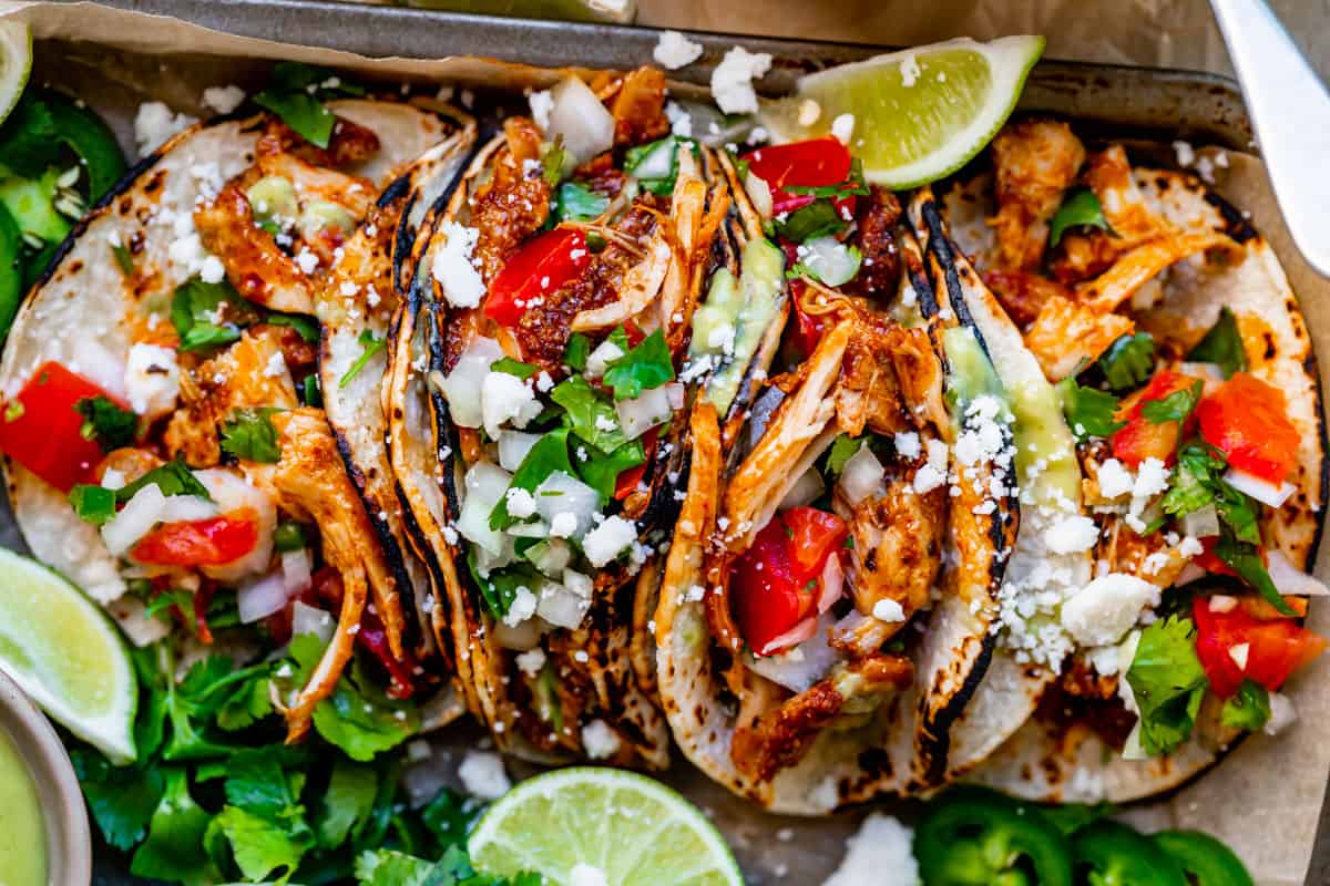 four tacos side by side with juicy chicken, tomato, onion, cilantro, and lime.