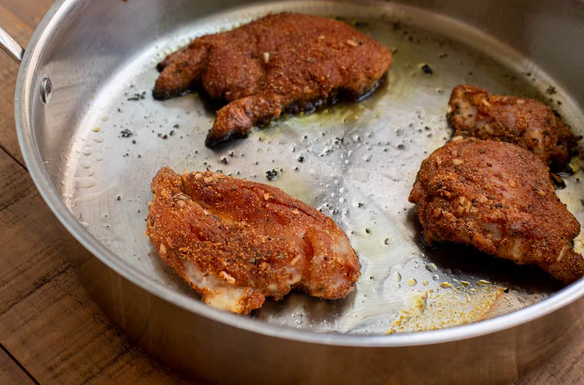 three well seasoned chicken thighs frying in a large saute pan on the stove.
