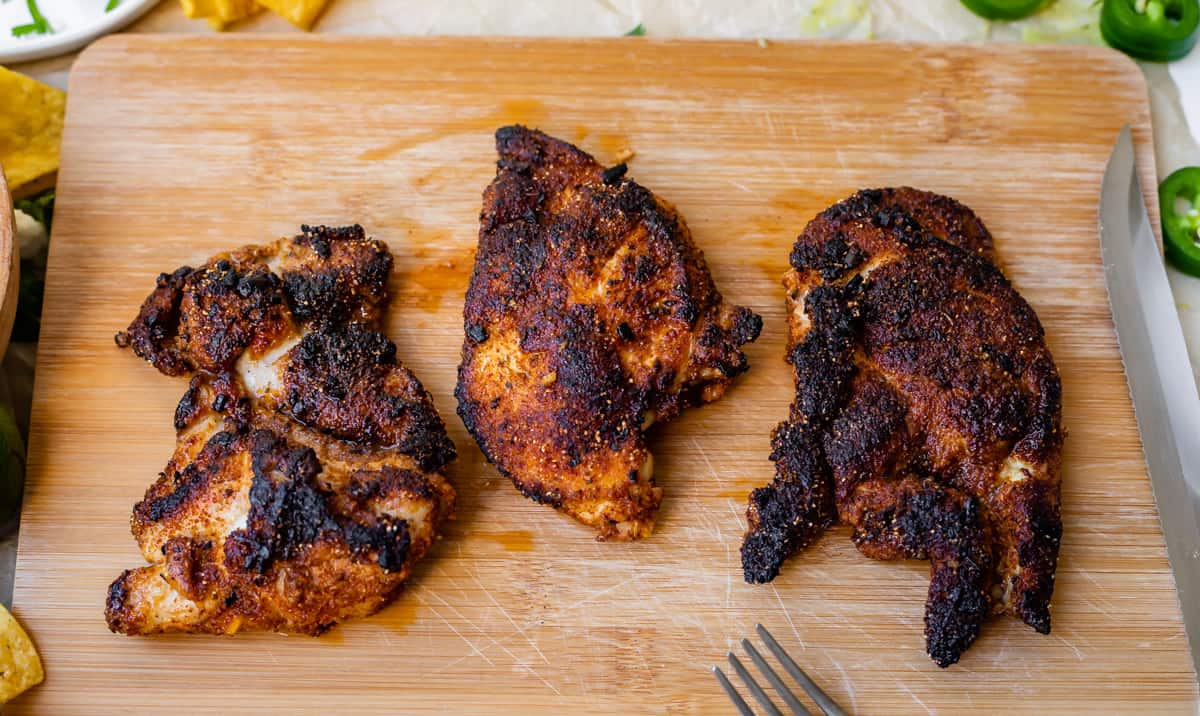 cutting board with three well seasoned and blackened chicken thighs ready to be cut.