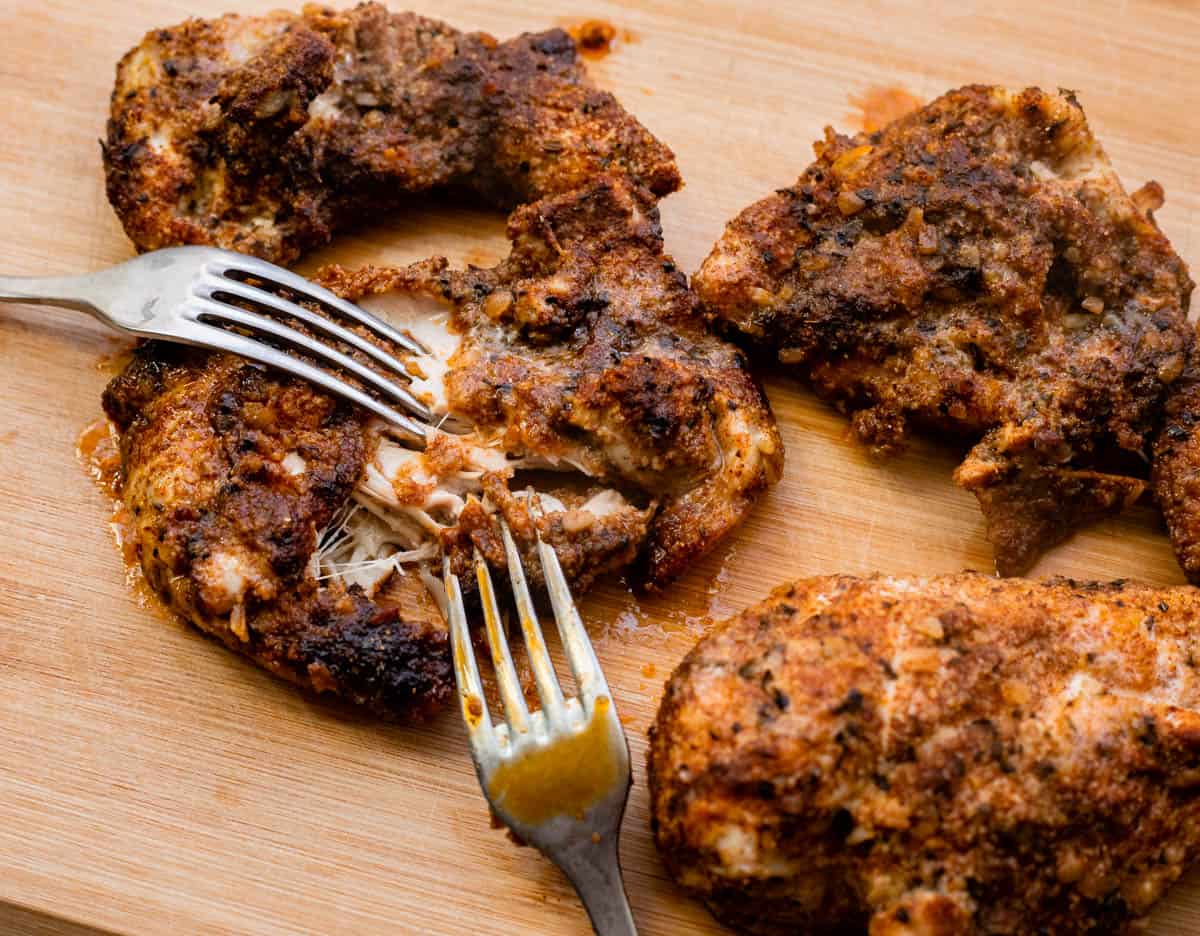 four pieces of chicken on a cutting board with two forks pulling into one piece to shred.