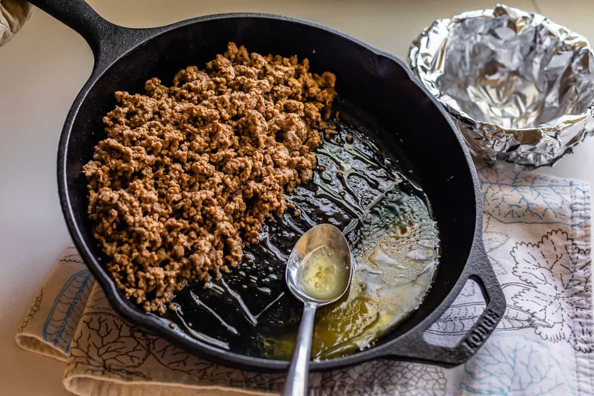draining grease from a cast iron skillet full of cooked ground beef.