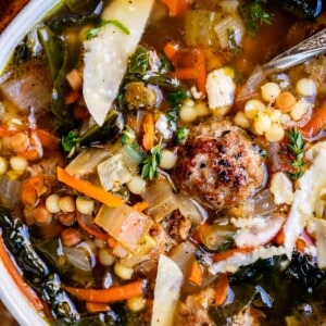close up of italian wedding soup with deep broth, meatballs, pasta and greens in a white bowl.