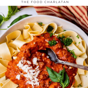 A bowl of wide pasta noodles topped with thick, chunky bolognese sauce, garnished with fresh basil and grated cheese. A spoon rests in the sauce, with a striped cloth and herbs nearby. Text reads “Bolognese Sauce.”.