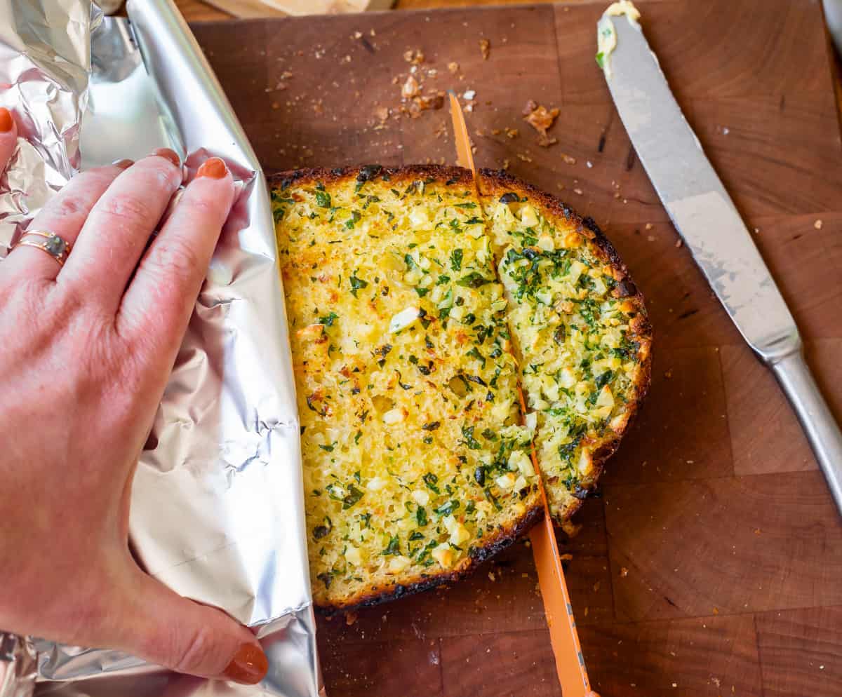 baked half of best garlic bread being sliced with a bread knife on a cutting board.
