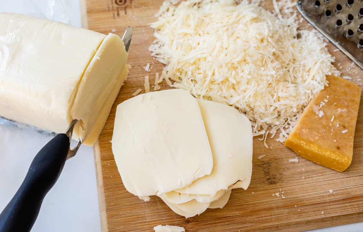 block of mozzarella cheese being sliced with a knife, shredded parmesan in the background.