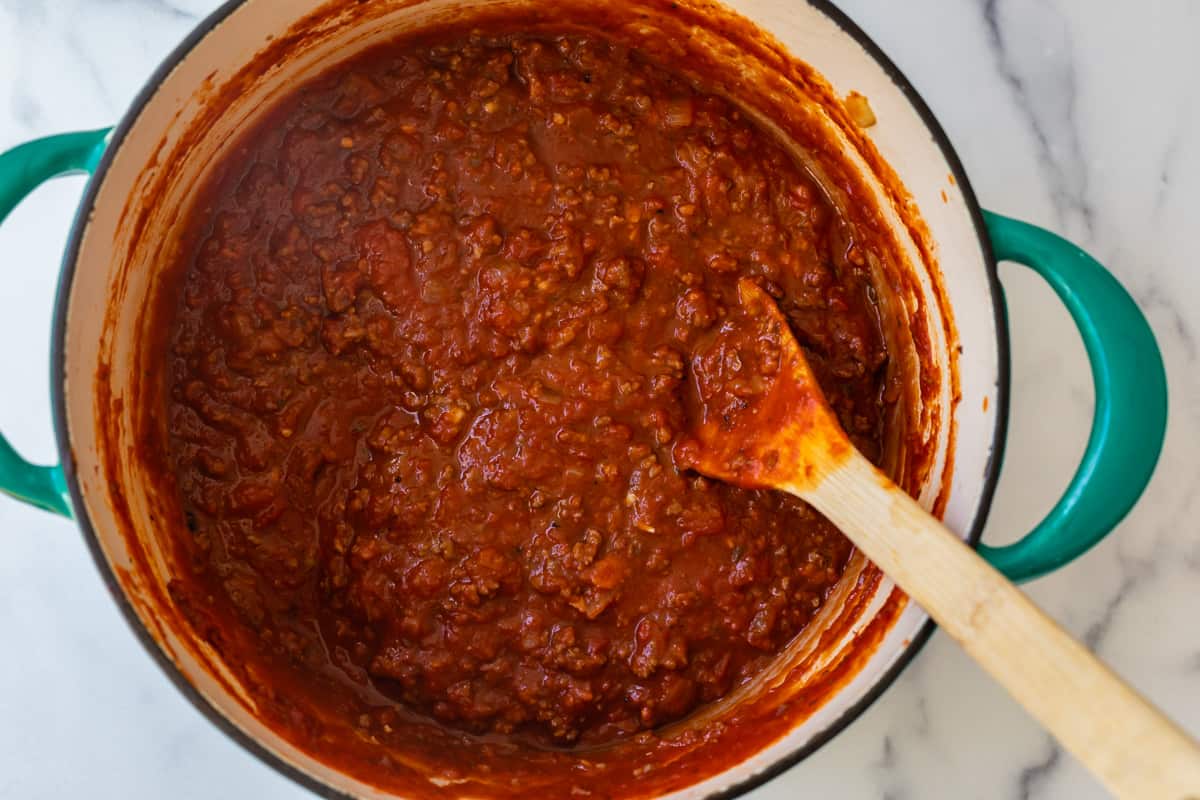 teal cast iron pot full of read meat sauce with a wooden spoon.