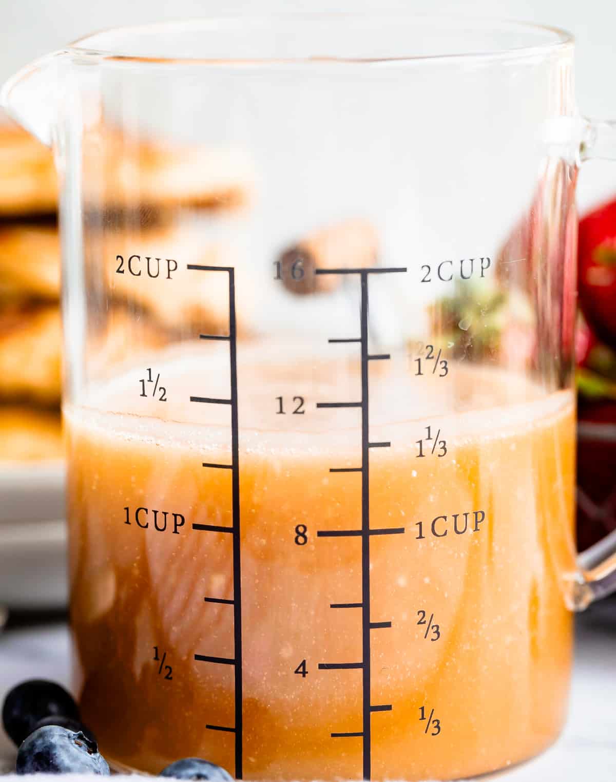 Buttermilk syrup in a glass measuring cup with blueberries in the foreground.