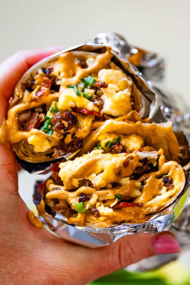 Two breakfast burrito wrapped in foil with sauce on top with bites taken out.