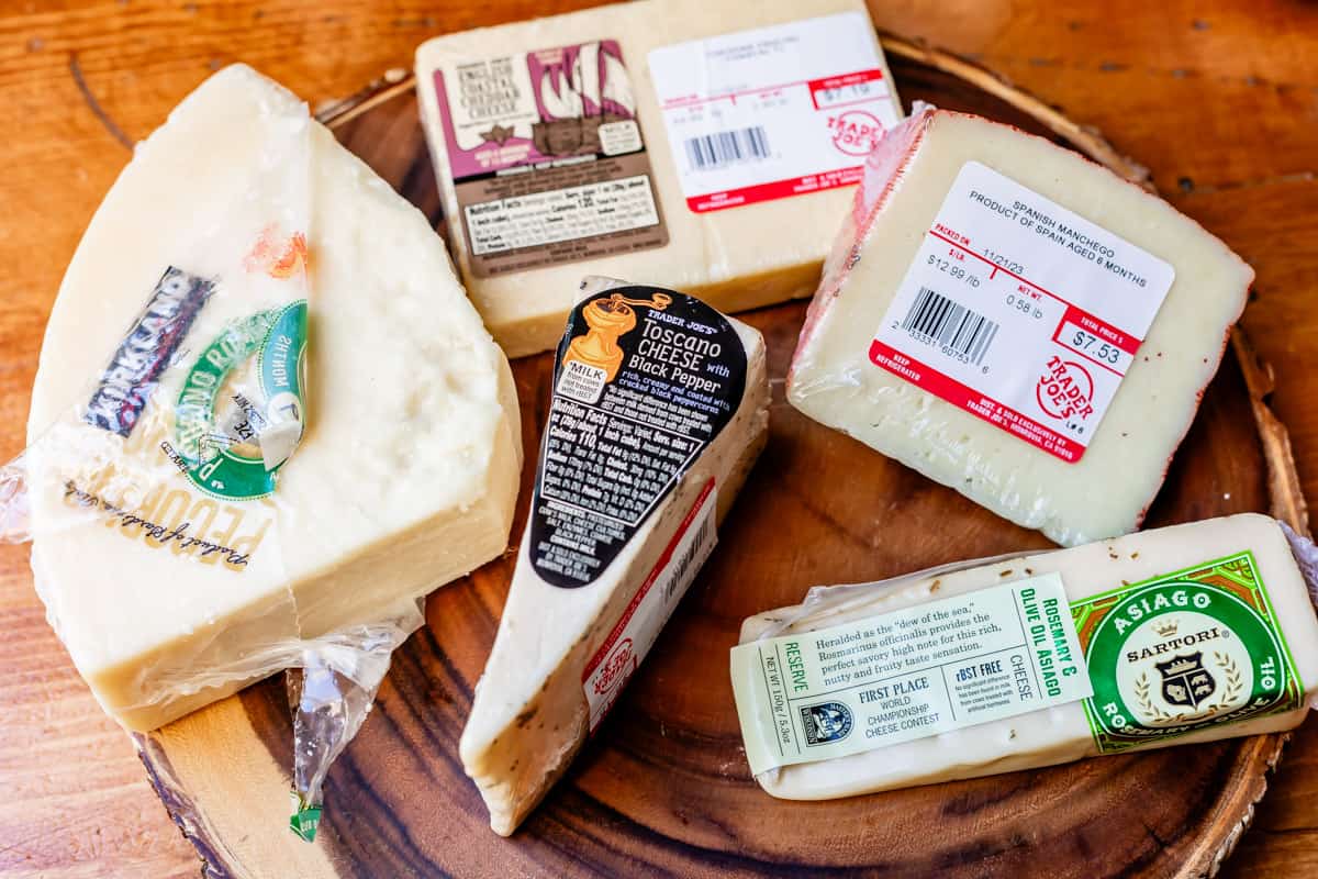Arrangement of hard cheese on wood board including pecorino romano, black pepper toscano, white cheddar, manchego, and asiago with rosemary.