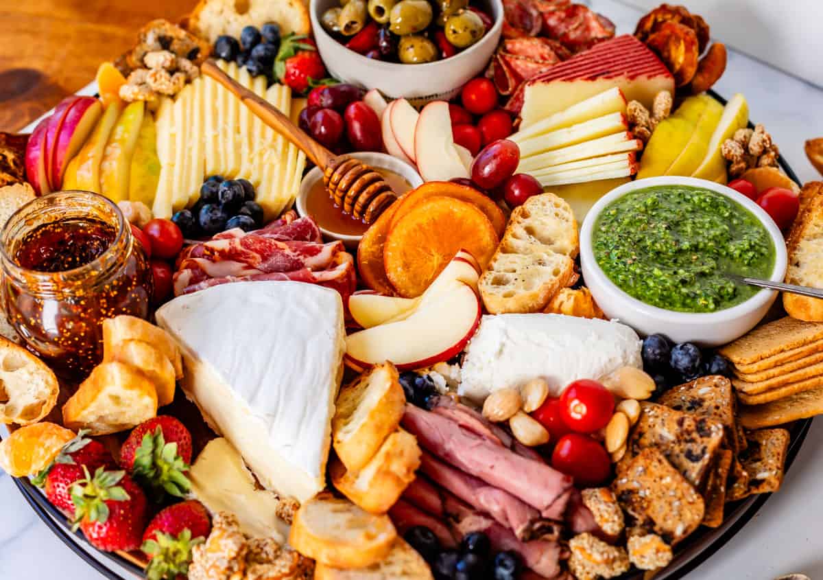 a round charcuterie board with apples, oranges, pesto, pastrami, nuts, etc.