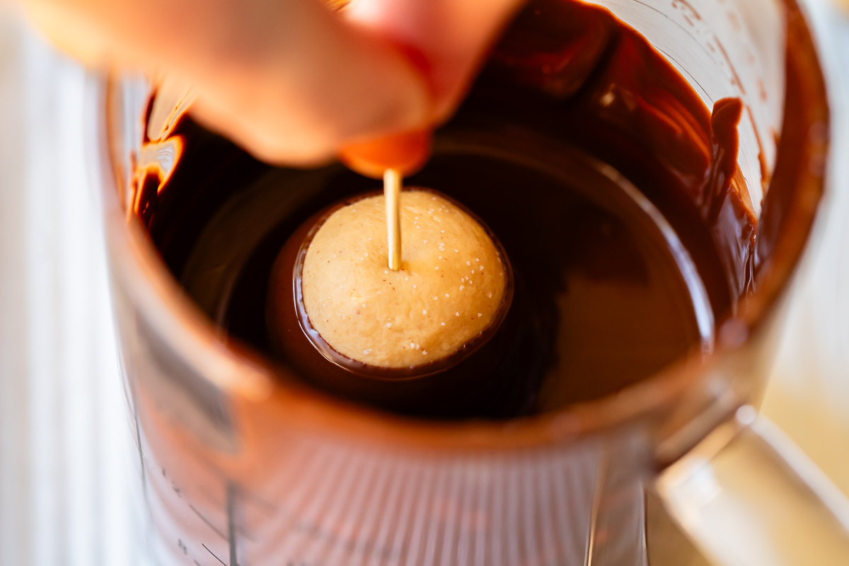 close up of fingers holding a toothpick in a peanut butter dough ball being dipped into chocolate.