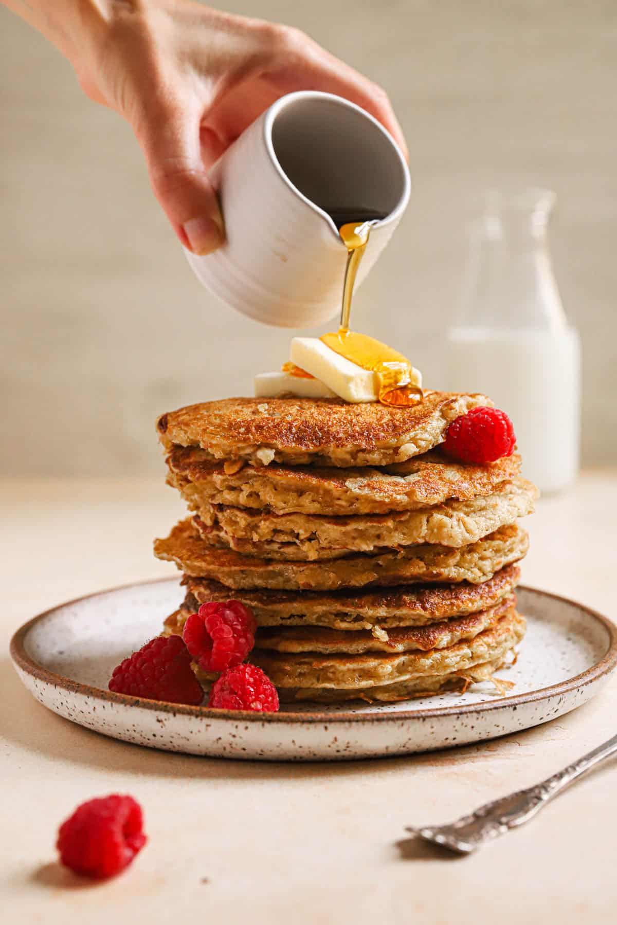 Pouring syrup on stack of pancakes with butter and raspberries.