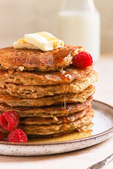 Short stack of oatmeal buttermilk pancakes with raspberries and syrup drizzling down on ceramic plate.