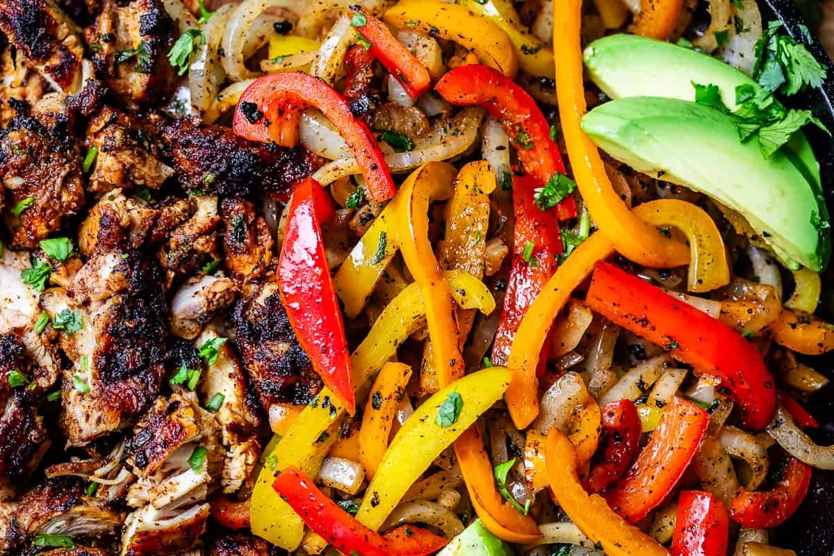close up of cooked chicken alongside sauteed onion and bell pepper, with avocado garnish.