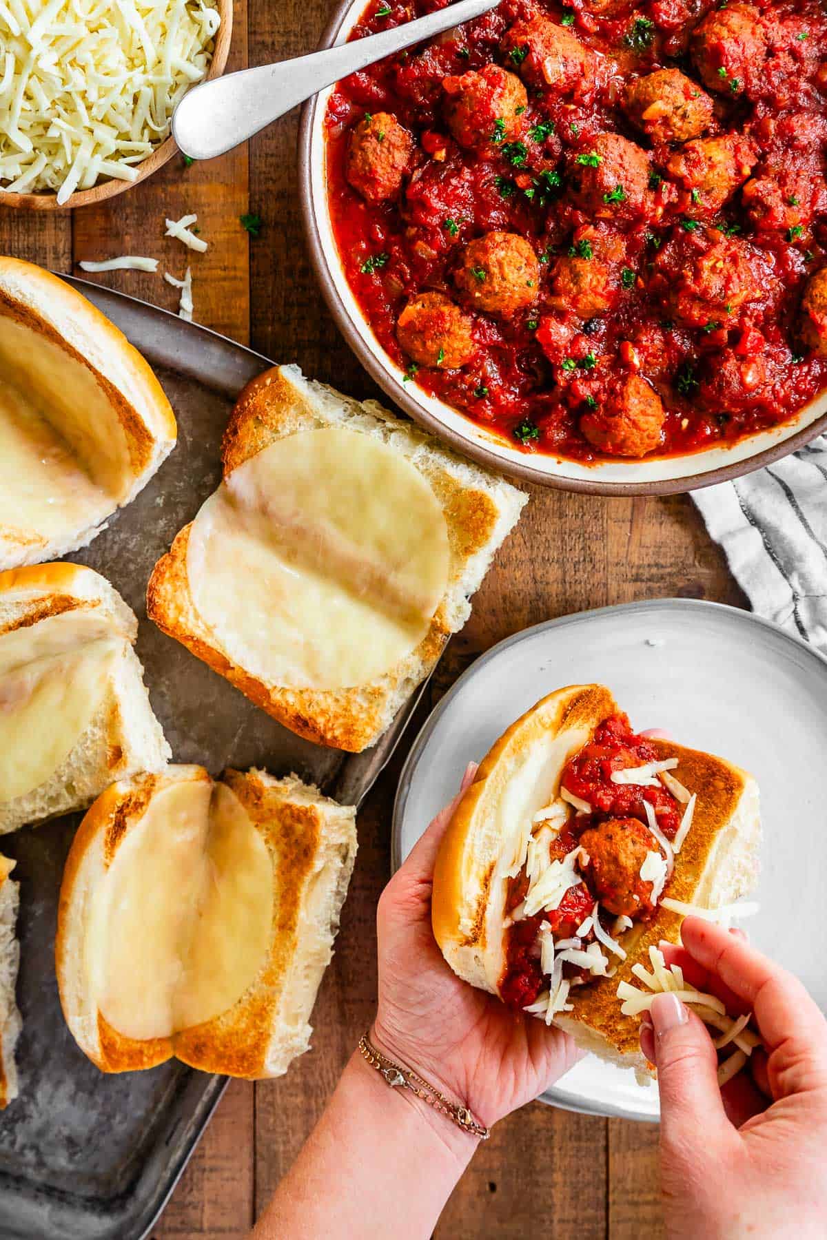 hands sprinkling cheese over meatballs and sauce in a toasted bun with melted cheese.