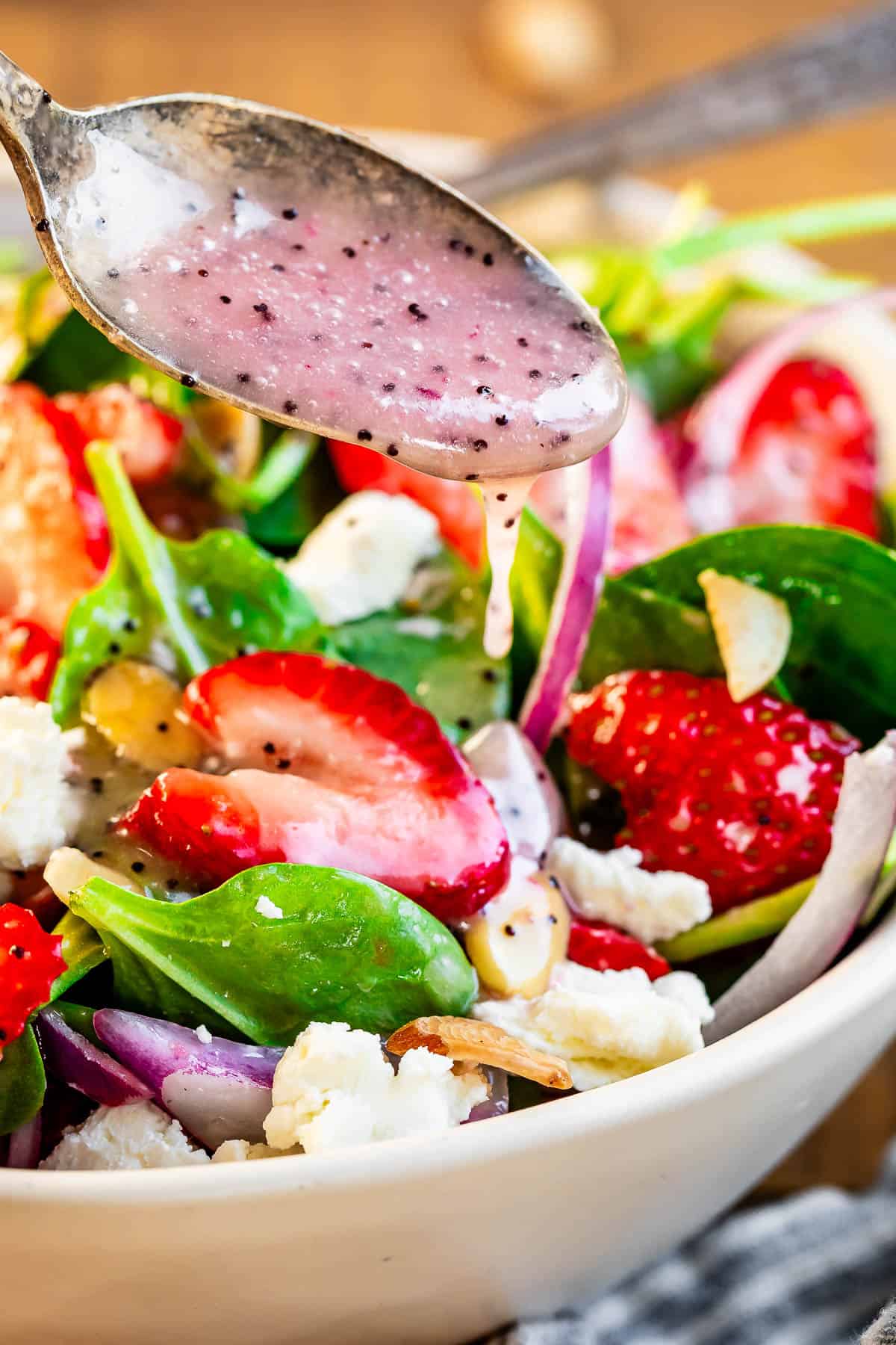 a metal spoon pouring poppyseed dressing over the top of spinach strawberry salad in a bowl.
