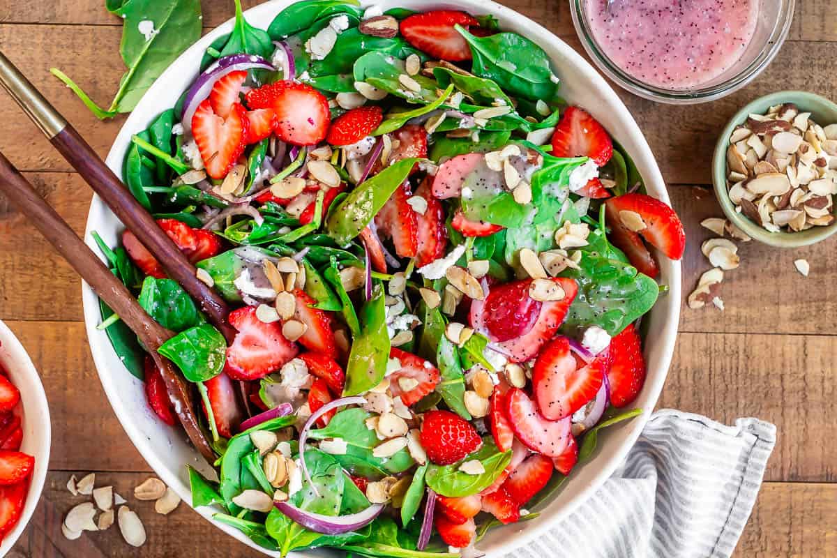 ceramic serving bowl filled with spinach strawberry salad with poppyseed dressing and salad tongs.
