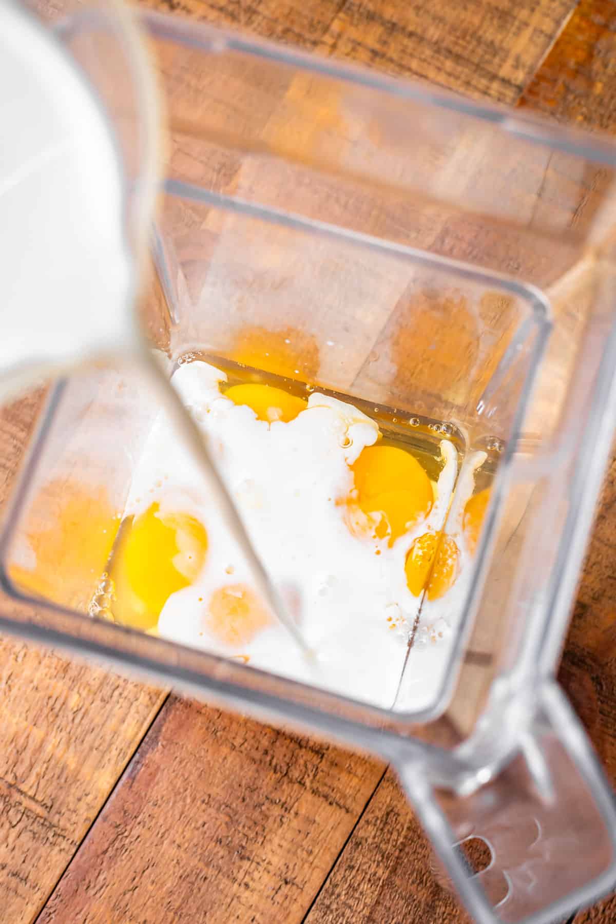 milk being poured from a measuring cup into a blender that already has eggs in it.