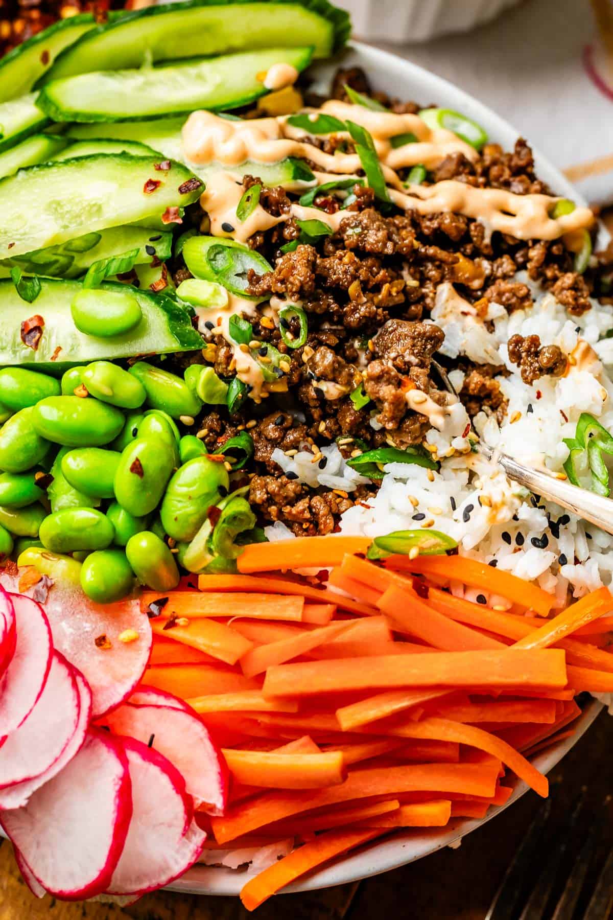 spoon digging into a bowl with rice, Korean beef, edamame, carrots sticks, cucumber, and more.