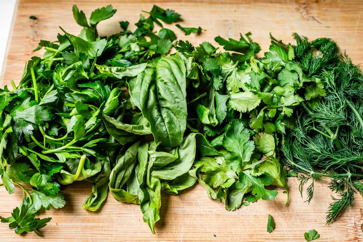 parsley, basil, cilantro, and dill lined up on a wooden cutting board to be chopped.
