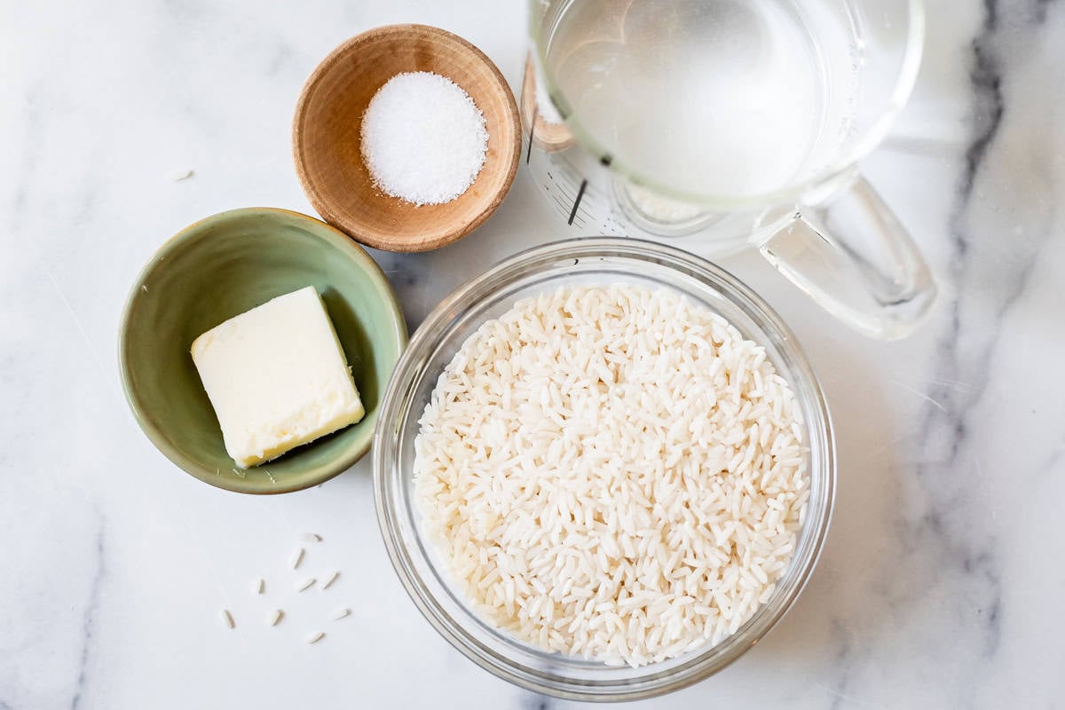 Measured ingredients for cooking rice in a pot including salt, butter, dry white rice, and water.