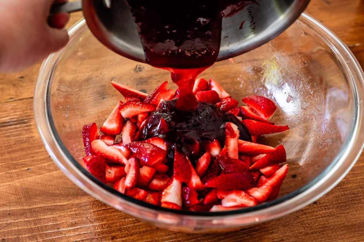 a hand pouring warmed jam from a metal pan into a bowl with fresh topped strawberries.
