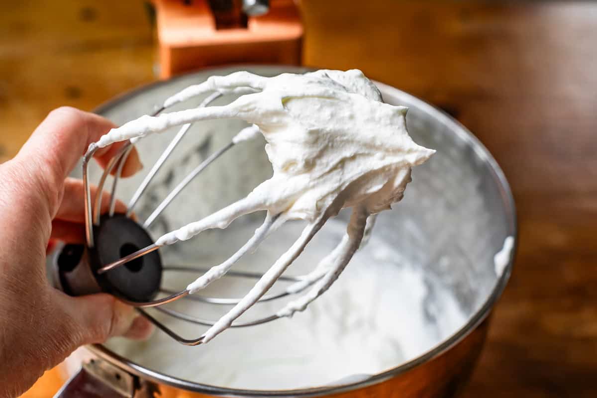 stiff whipped cream on whish attachment with metal mixing bowl with whipped cream in background.