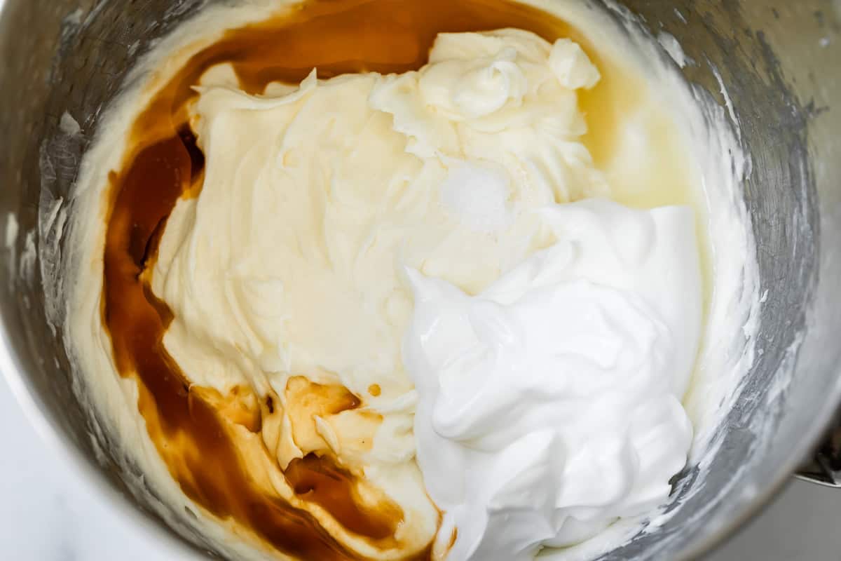 whipped cream cheese in metal mixing bowl with vanilla, sour cream, and sugar piled on top.