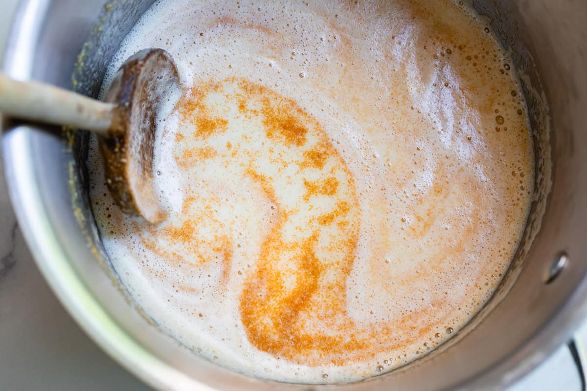 wooden spoon stirring through browned butter with foam on top in metal pan.