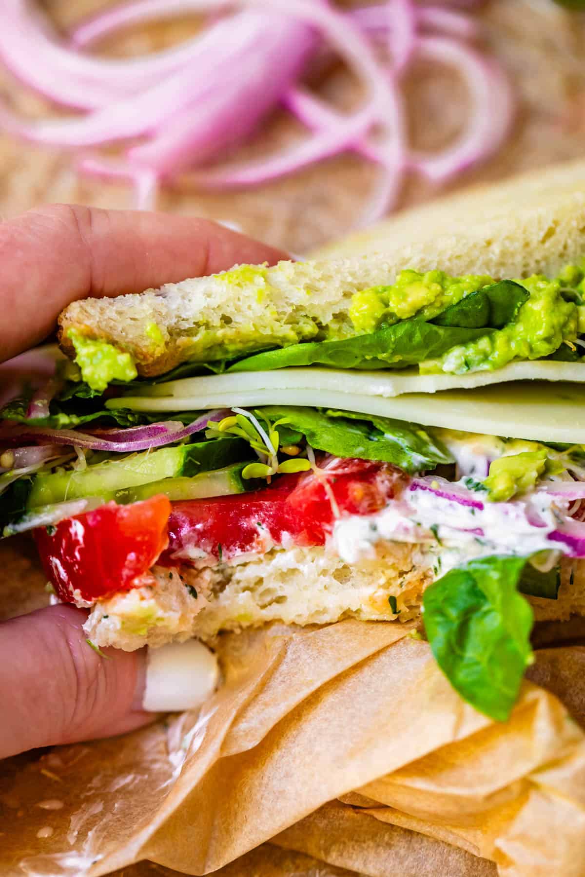 a hand holding a fresh veggie sandwich stuffed with spread, tomato, cucumber, and more.