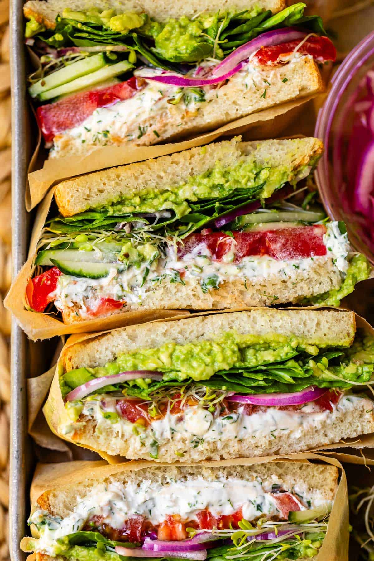 a metal baking tray filled with sideways stacked fresh vegetable sandwiches wrapped in parchment.