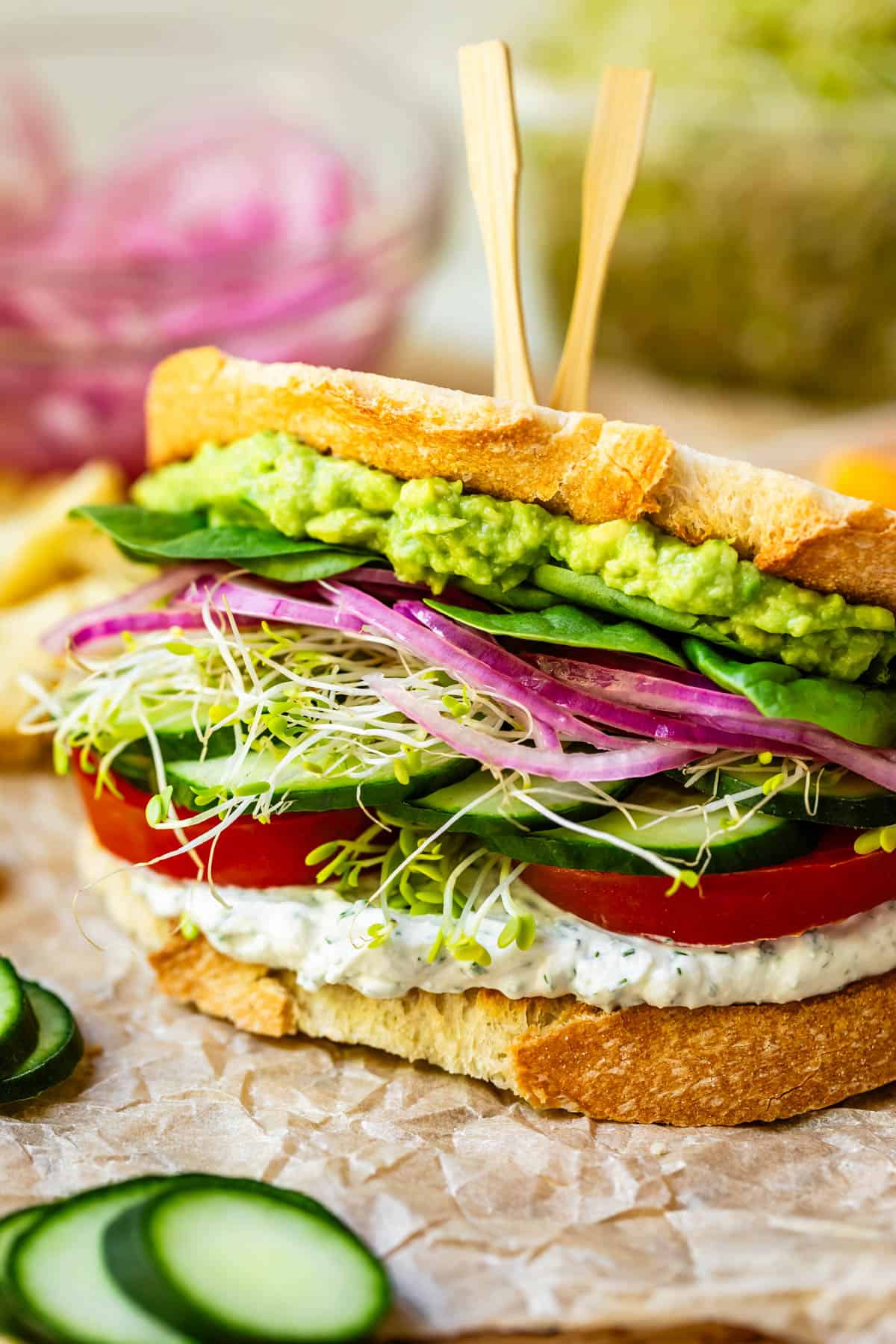 a delicious fresh veggie sandwich with 2 bamboo sandwich skewers in the top on parchment paper.