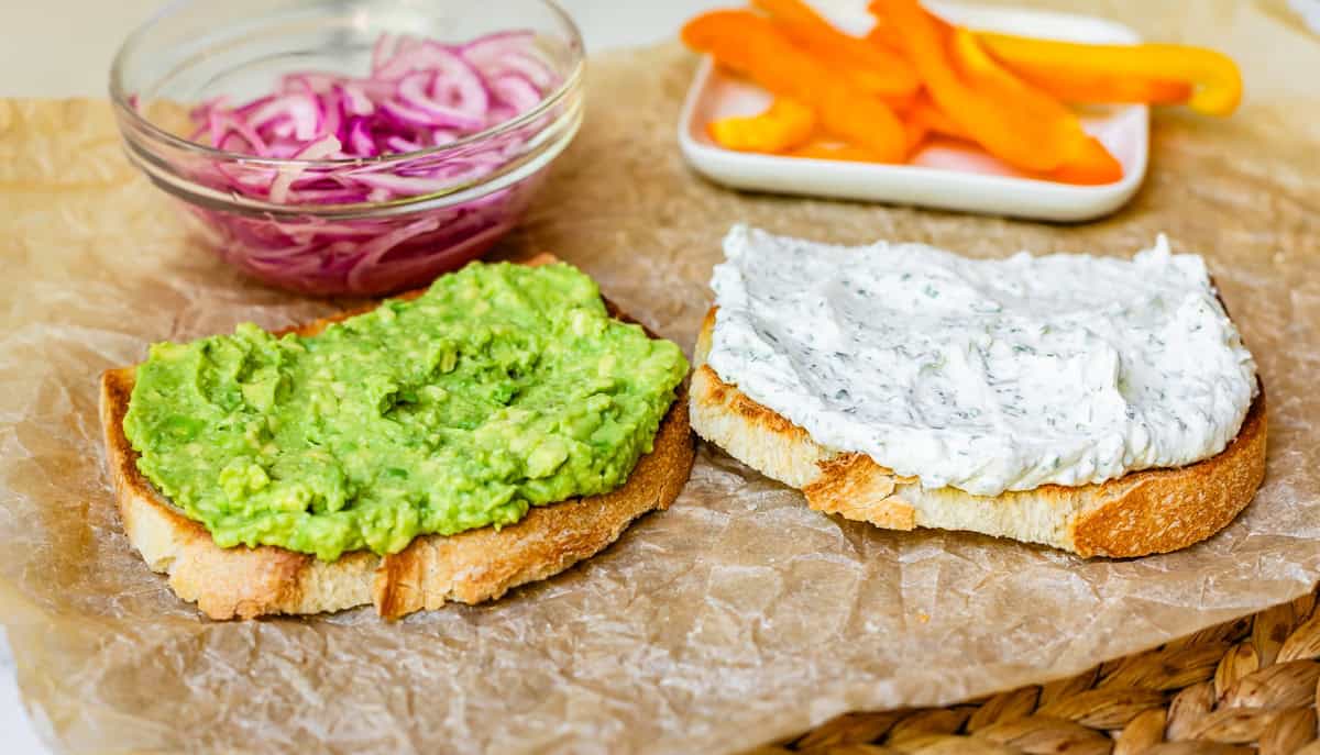 two slices of bread with cream cheese spread on one and mashed avocado on the other.