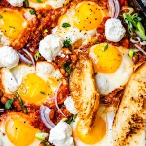 cast iron skillet with shakshuka, a piece of toast stuck into it, and red and green onion garnish.