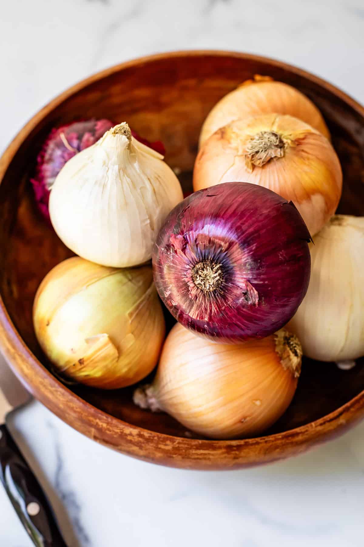 wooden bowl filled with different onions, on a countertop next to a knife.