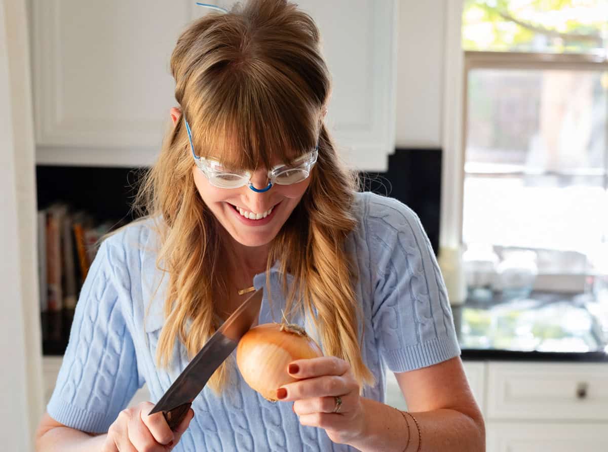 woman wearing swim goggles using the back of a chefs knife to peel skin off an onion.