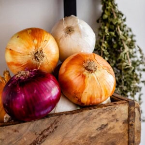 red, white, and yellow onions stacked in a basket on the wall with dried thyme hanging next to it.