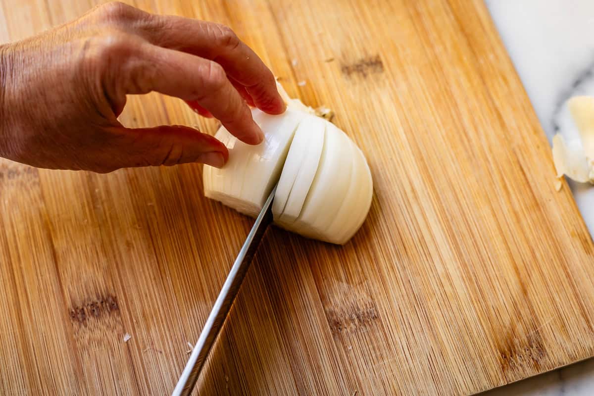 Cutting vertical cuts off of root of an onion with a chef's knife on bamboo cutting board.