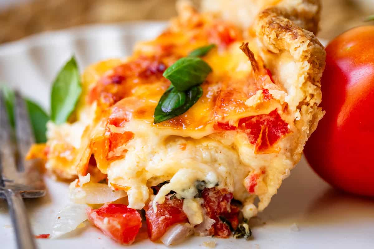 Side view of cheesy tomato pie with bite taken out of it on white plate by a tomato.