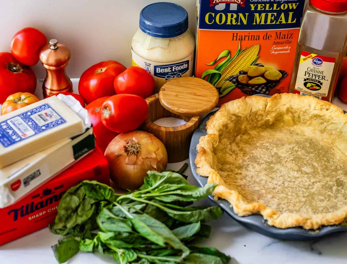 Ingredients laid out ready to make tomato pie including tomatoes, salt, mayonnaise, corn meal, cayenne pepper, cheese, greens, onion, and a pie crust.