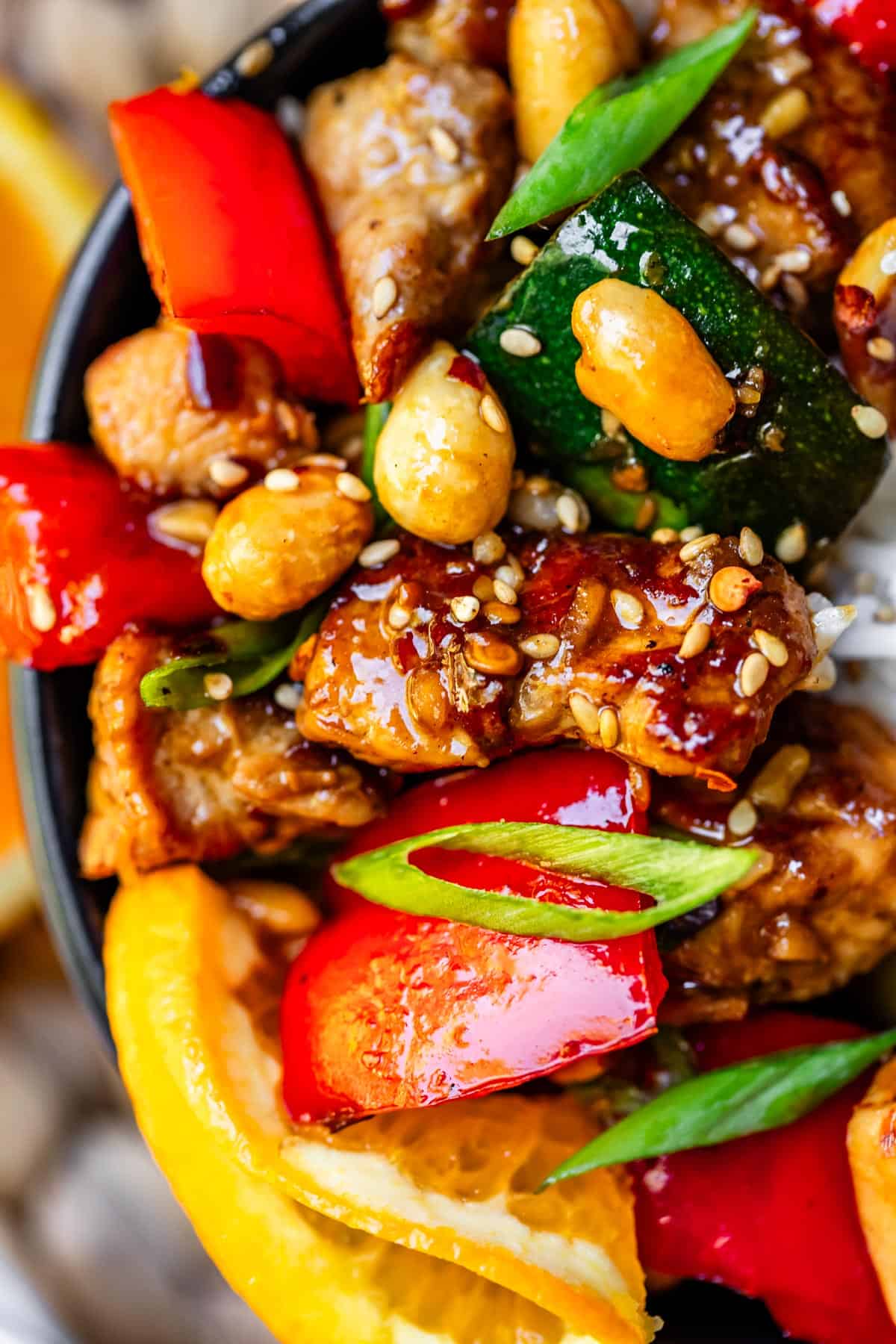 Close up of kung pao chicken in black ceramic bowl with bright red and green veggies and chicken covered in sauce and sesame seeds.